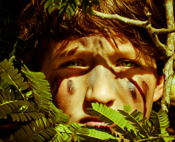 Photo of a young boy, his face painted with camouflage, staring from out of a bush.
