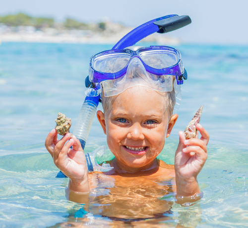 Young child swimming in the sea with goggles on his head holding shells